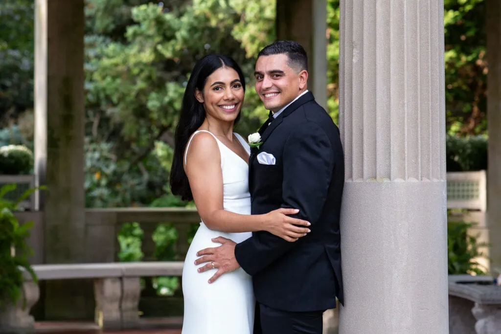 A bride and groom with arms around each other smiling next to a stone pillar