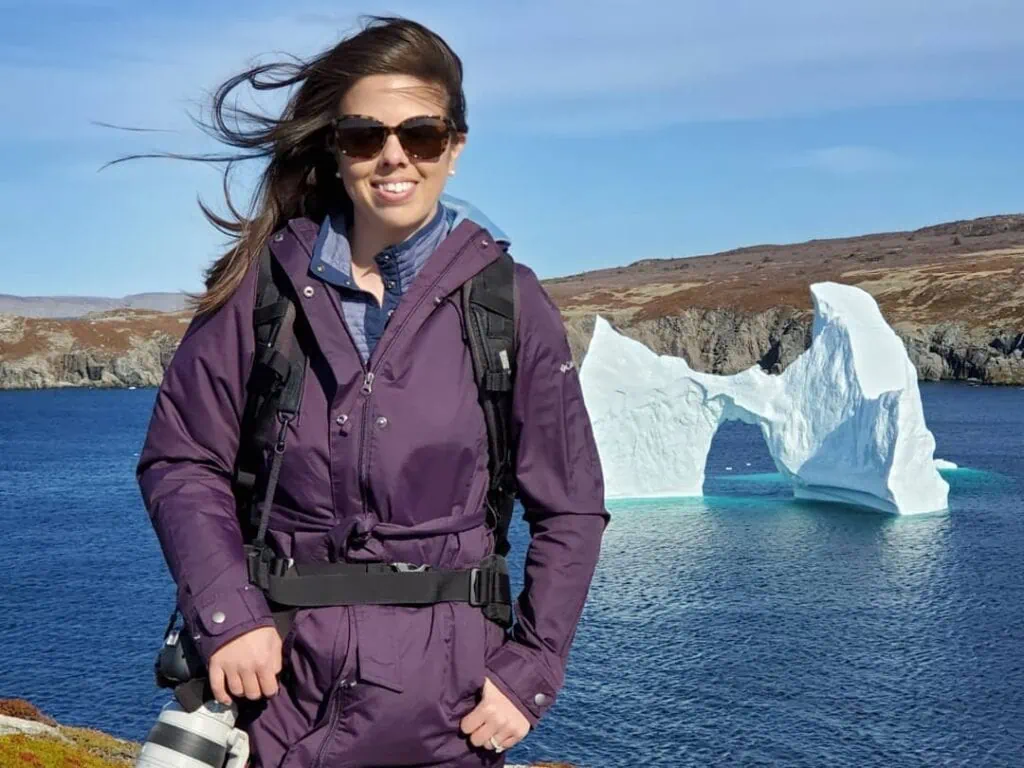 A woman with purple jacket and hair blowing in the wind holding a camera with an iceberg in the ocean behind her