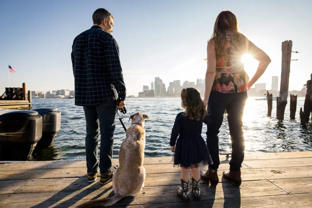 A mom and dad stand on a dock looking at the city of boston while holding their daughters hand and their dog on a leash
