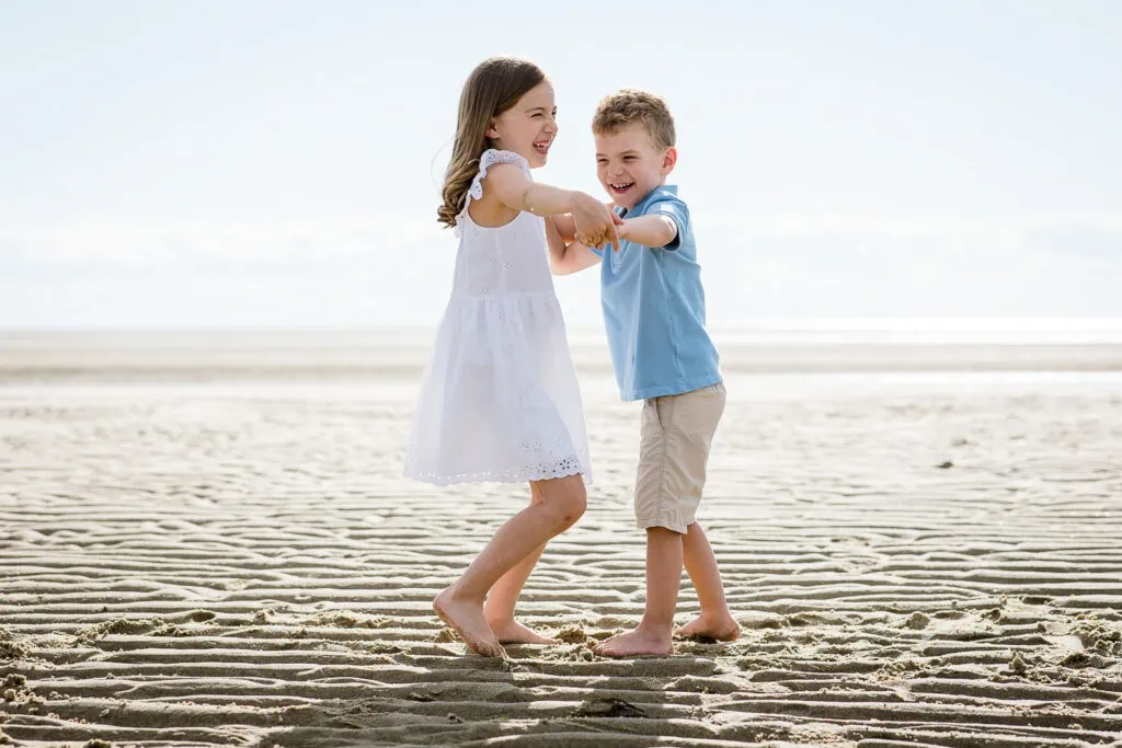 A little girl in white and a little boy in blue hold hands and dance at the beach