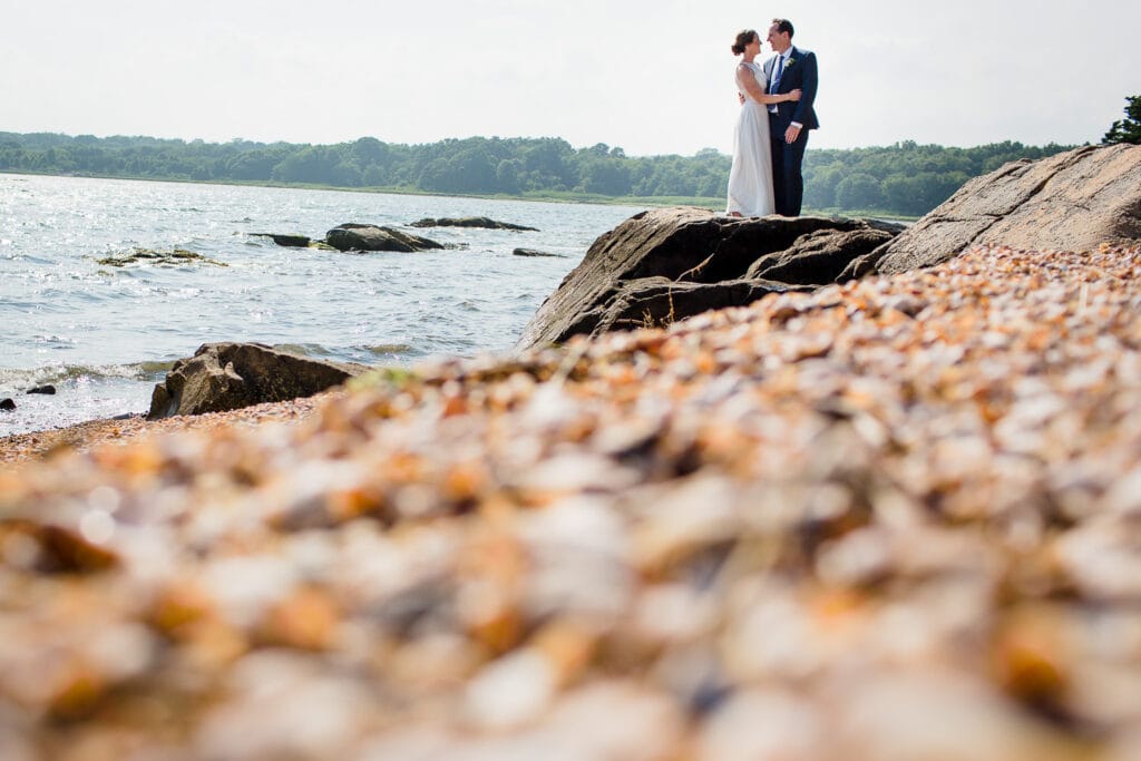 A foreground of beach pebbles leads to a bride and groom standing on a rocky outcropping next to the ocean
