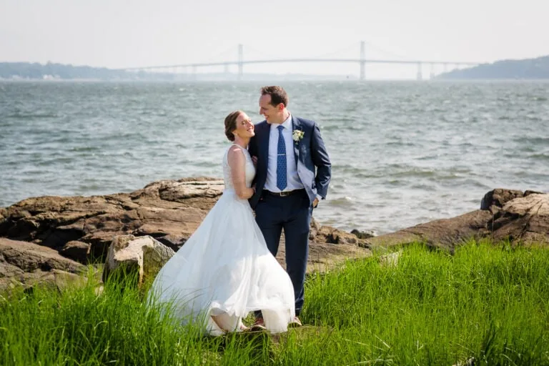 A bride and groom stand in the grass near the edge of the ocean with the mount hope bridge behind them