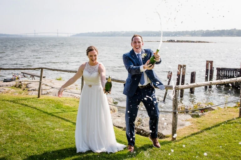 A bride and groom spray champagne at their Mount Hope Farm wedding near the cove cabin with the ocean and mount hope bridge in the background