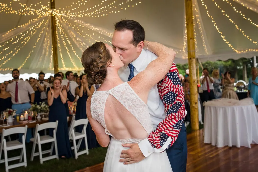 A newlywed couple kissing on the dance floor under a tent filled with string lights