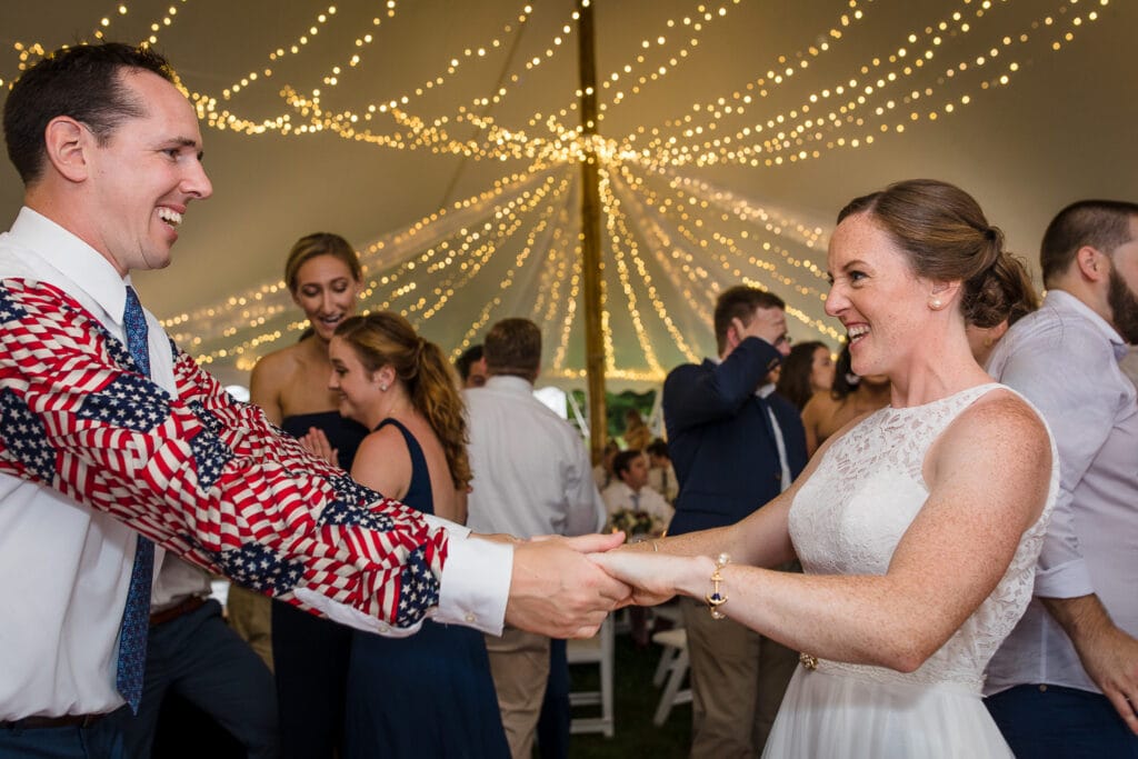 A bride in a white lace dress and groom in a white dress shirt with american flag patterned arms dancing with outstretched arms holding each other under at tent at their Mount Hope Farm wedding