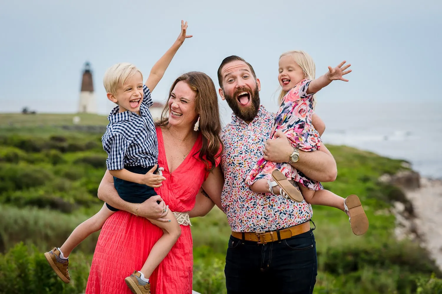 A man and woman hold two children as they laugh and stick tongues out near the Pt Judith Lighthouse