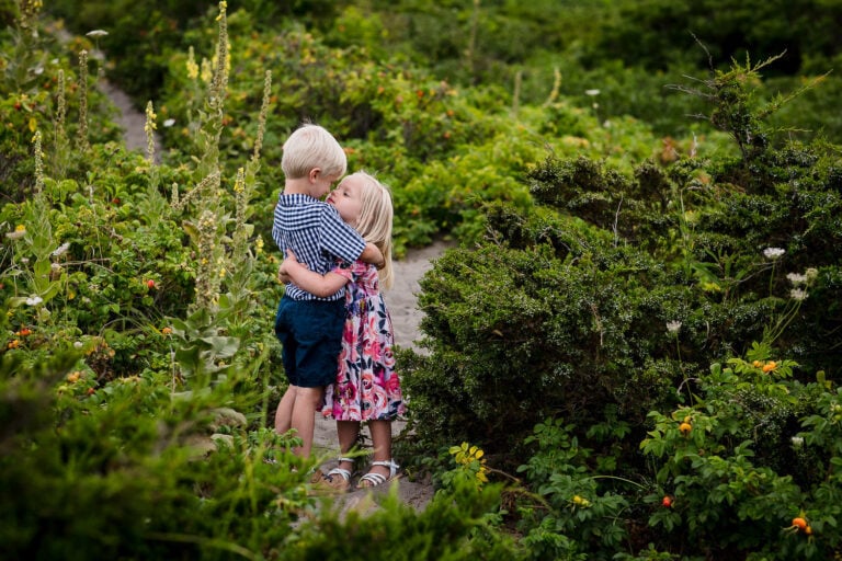 A little blonde girl and boy hug and touch noses on a path through beach roses at a rhode island family photography session