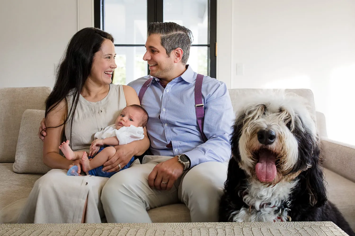A man and woman sit on a couch holding a baby as their large dog looks at the camera