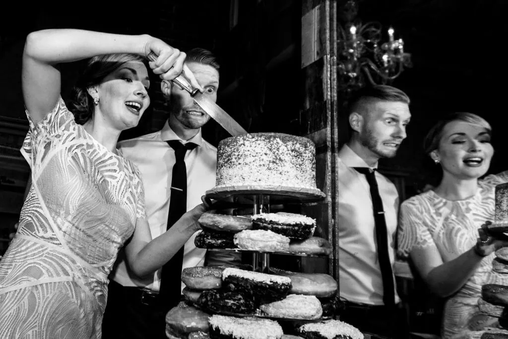 A bride cuts a wedding cake as the groom winces and they are both reflected in a mirror