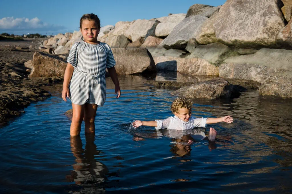 A little girl standing in a tidal pool next to a little boy who is sitting in the pool