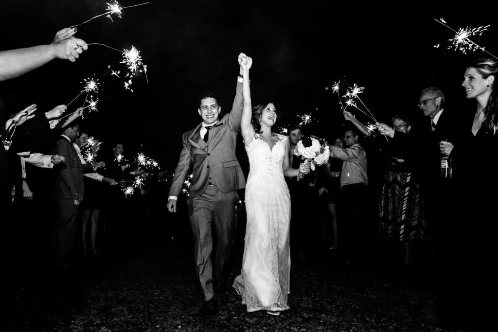 A newlywed couple run with holding hands raised above their heads through a tunnel of sparklers