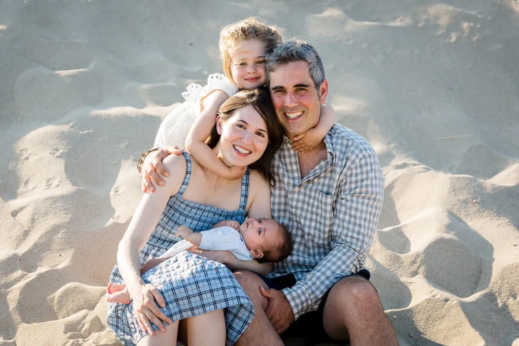 A mom and dad sitting in the sand holding a newborn with a little girl hugging them from behind