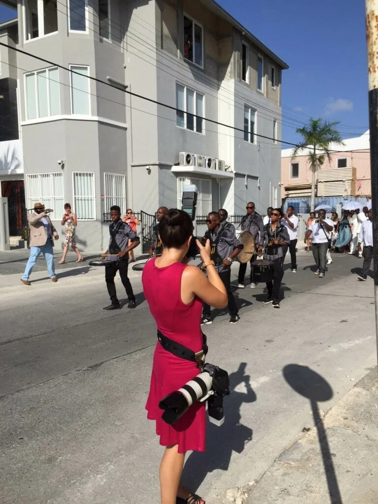A woman wearing a red dress with one camera up to her eye and another on her waist taking photos of a band marching down a street