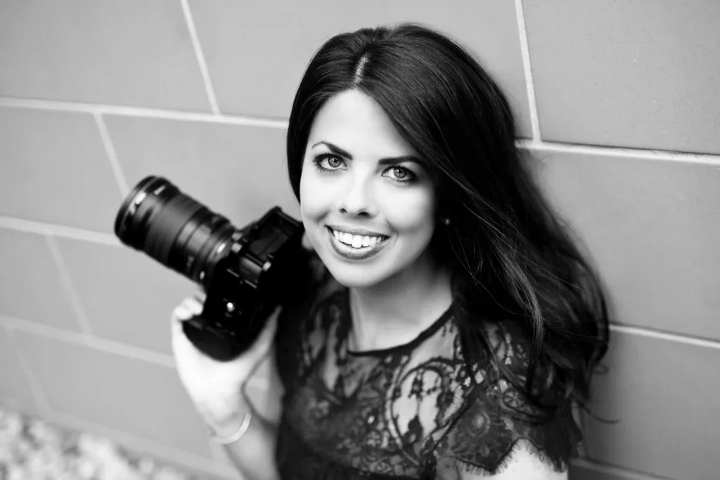 Woman with dark hair smiling and leaning up against a wall with a professional camera in her hand