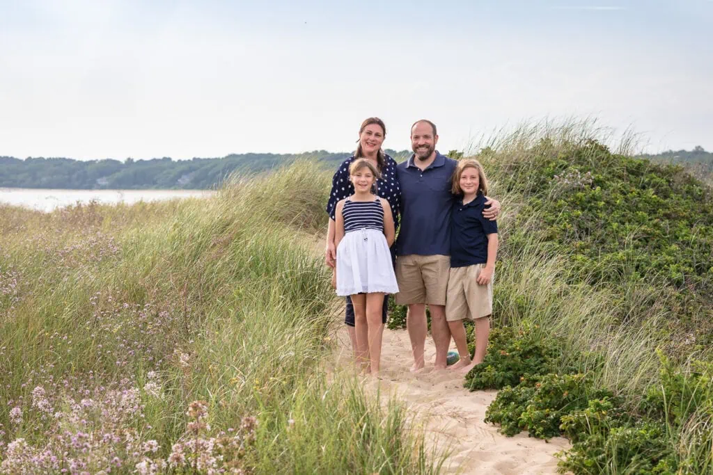 A family of 4 wearing white and navy stand in a small sand dune by the ocean