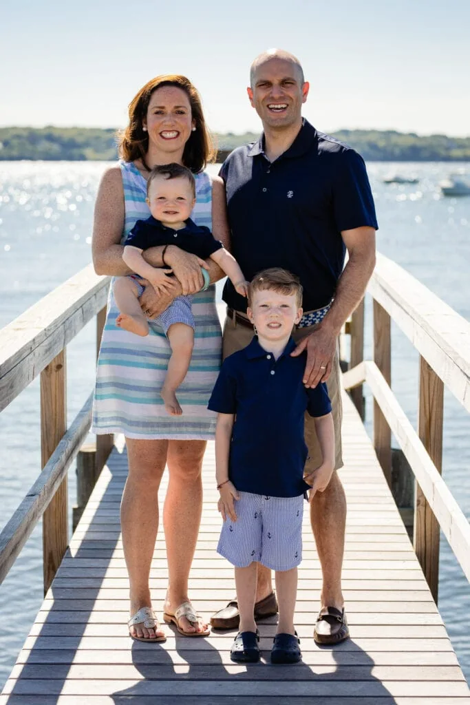 A mom, dad, and two little boys pose on a dock by the ocean