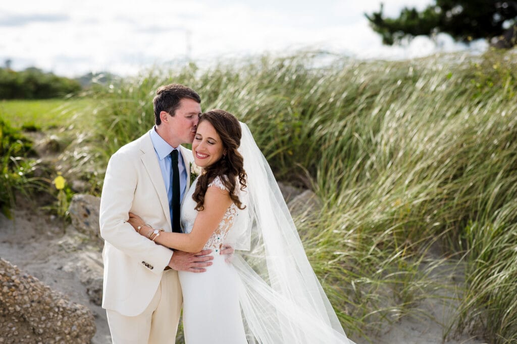A groom wearing a tan suit kisses the forehead of his bride in white dress and veil in front of beach grasses