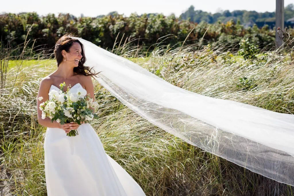 A bride in white dress and long flowing veil looks off to the side in front of beach grasses