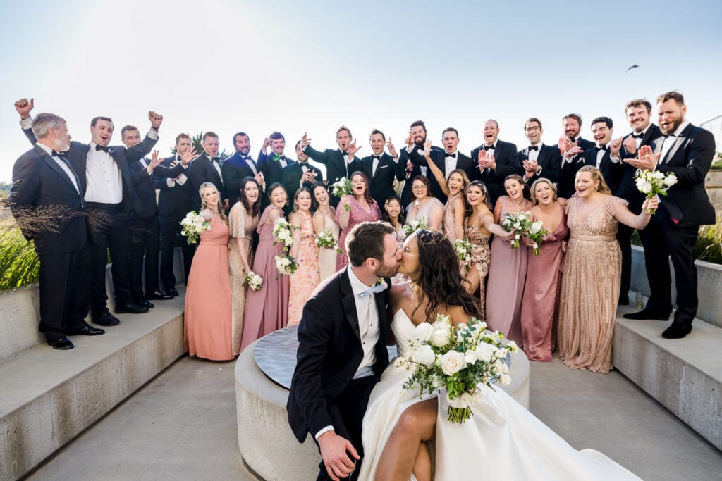 A bride and groom kiss sitting on a firepit with a cheering massive bridal party behind them