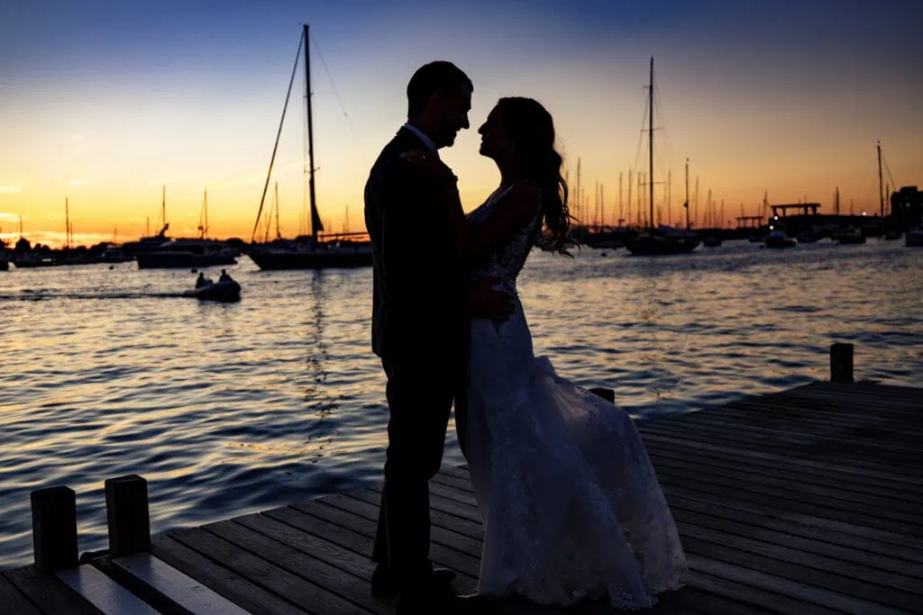 A silhouette of a bride and groom standing on a dock with boats in the background and a sunset sky at The Bohlin Weddings and Events
