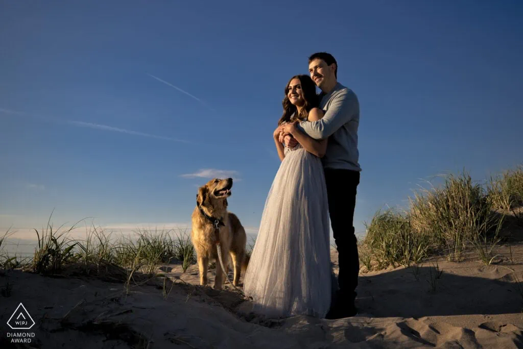 A man and woman embrace on a dune as their golden retriever dog looks up at them
