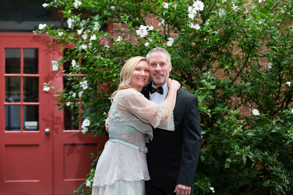 A couple hugging by a flower bush and red doors