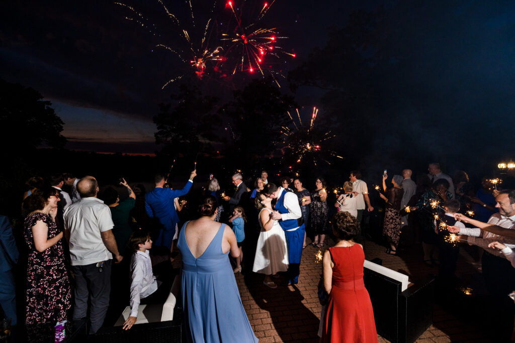 Fireworks go off above a patio filled with people surrounding a bride and groom kissing