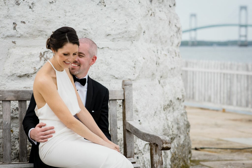 A bride sitting on her grooms lap by the newport harbor lighthouse and the newport bridge