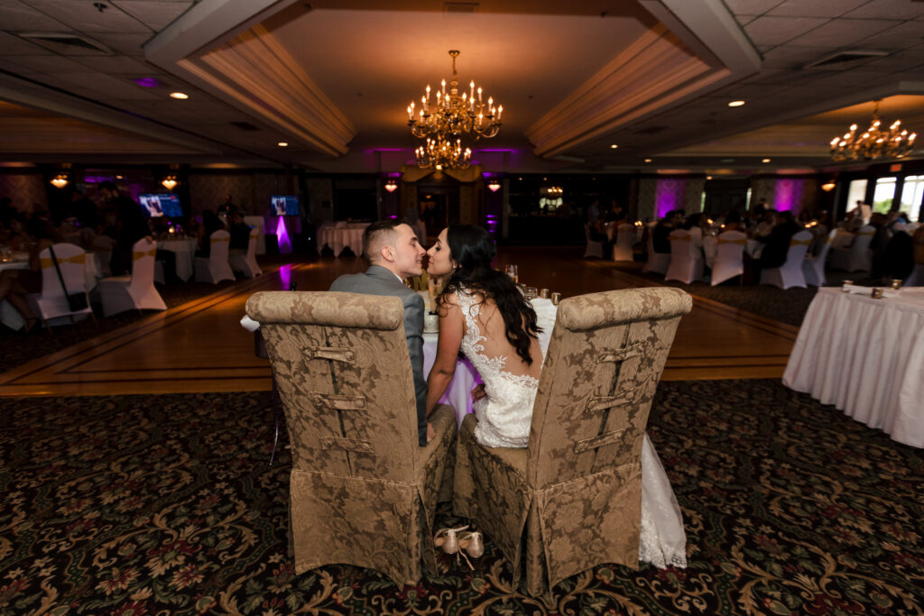 A bride and groom kiss while sitting in chairs at their sweetheart table