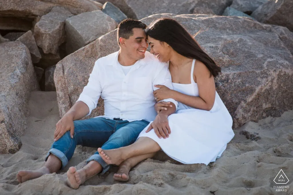 A couple wearing white sitting on the beach laughing with each other